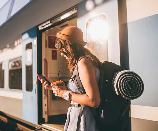 Жена с телефон в ръка и раница стои на гарата.