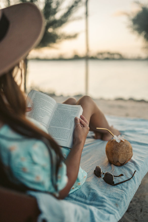 Жена със слънчева шапка, лежаща на плажа и четеща книга, кокосова напитка