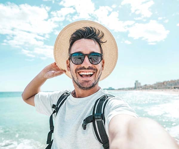 Млад мъж с шапка и слънчеви очила се усмихва на плажа