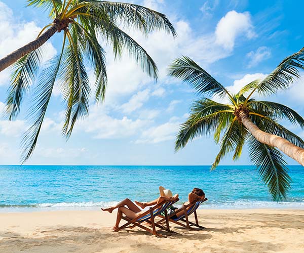 Жена и мъж лежат на плажа на шезлонги, заобиколени от палми.